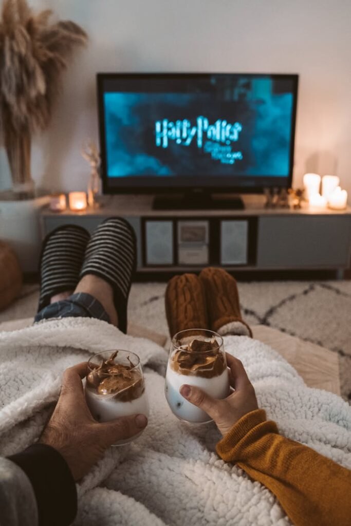 A couple enjoys a cozy movie night with drinks, wrapped in blankets, watching a Harry Potter film on TV.