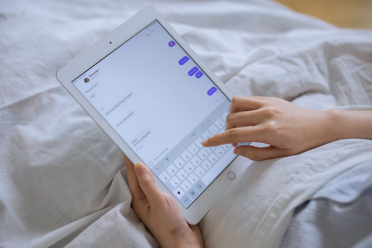 Close-up of hands typing on a digital tablet with messaging app in bed.